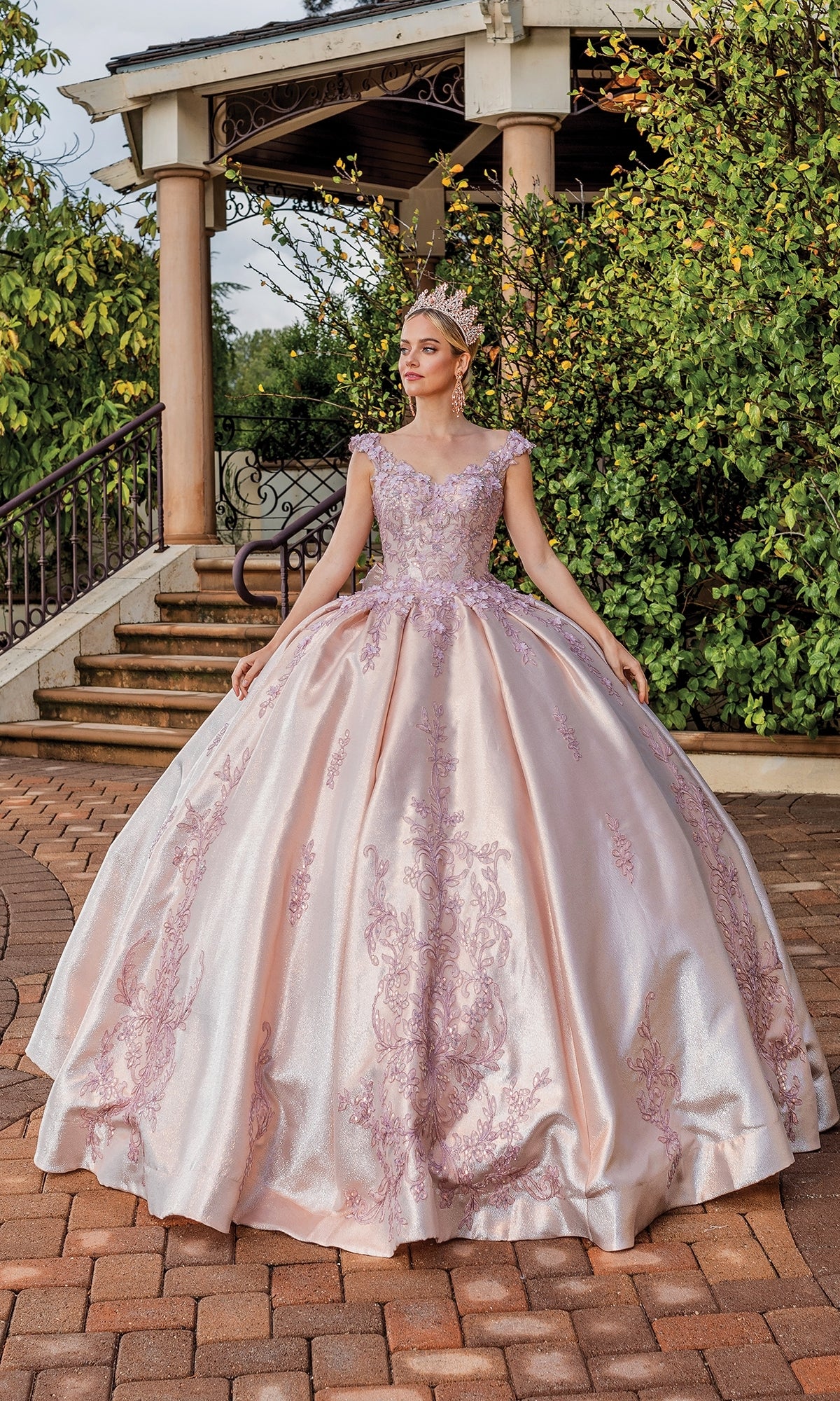 A woman in the Quinceanera Dress 1762 by Dancing Queen, a pink ball gown with floral embroidery and a tiara, stands on a brick path before stairs and greenery.